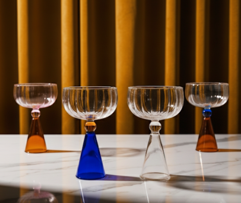 Set of four glass goblets with colored stems on a reflective surface with a striped background