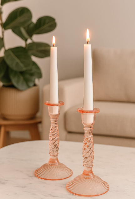 Two white candles in pink candlesticks on a table with a plant and sofa in the background.